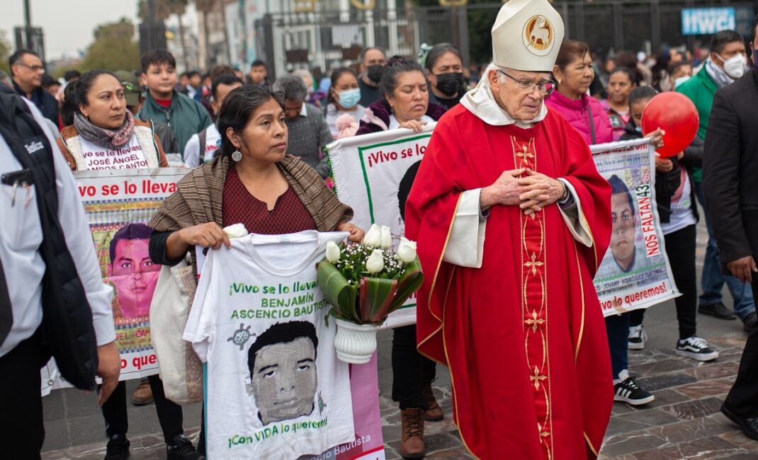 A crowd of parents, walk through the street with a bishop, holding images of their missing children