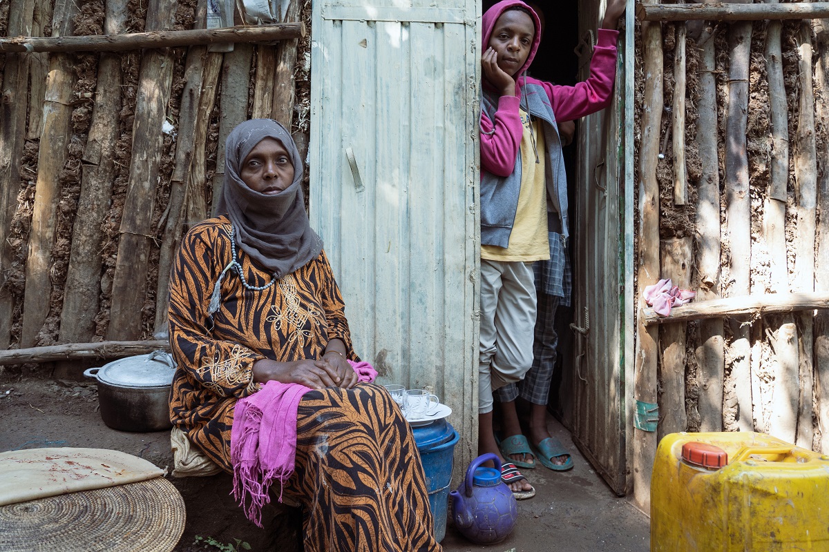  Photograph by Jaclynn Ashly showing Muntaha Suleiman, an Ethiopian woman in a black and orange stripped dress and a hijab, sitting outside her home in Gondar. Two other people are standing at the door and looking at the camera.