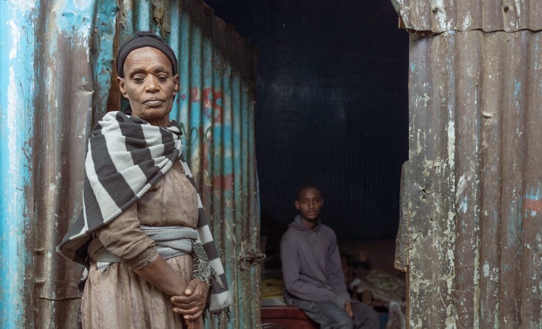  Photograph by Jaclynn Ashly showing an Ethiopian women in front of her shack house, looking at the ground. The door is open, in the house her son is sitting on the bed, looking at the camera.