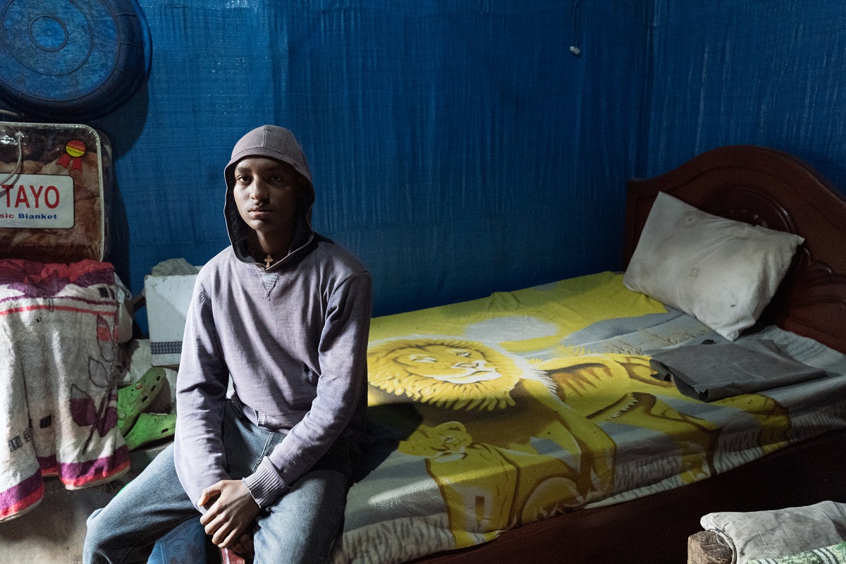 Photograph by Jaclynn Ashly showing a young Ethiopian man sitting on his bed looking at the camera, wearing a hood