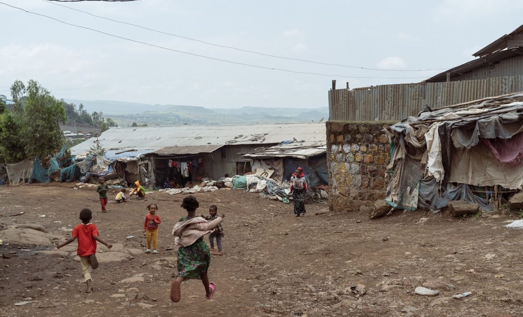 A group of children are running in the open, a woman with a child on her back walks towards the group. A tin building is surrounded by hanging clothes, sheets and nets:  Photograph by Jaclynn Ashly for Lacuna story on religious tensions in Ethiopia.