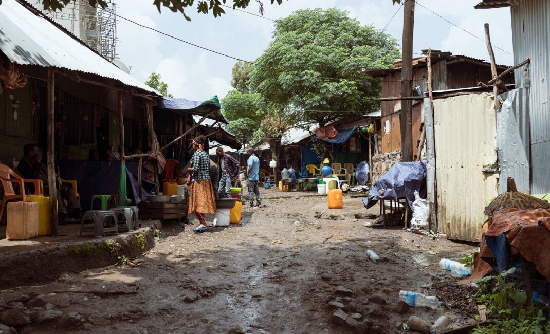 A neighbourhood in Gondar with a muddy road, tin buildings and people sitting and meandering around. A powerline cuts across the street and plastic bottles litter the ground:  Photograph by Jaclynn Ashly for Lacuna story on religious tensions in Ethiopia.