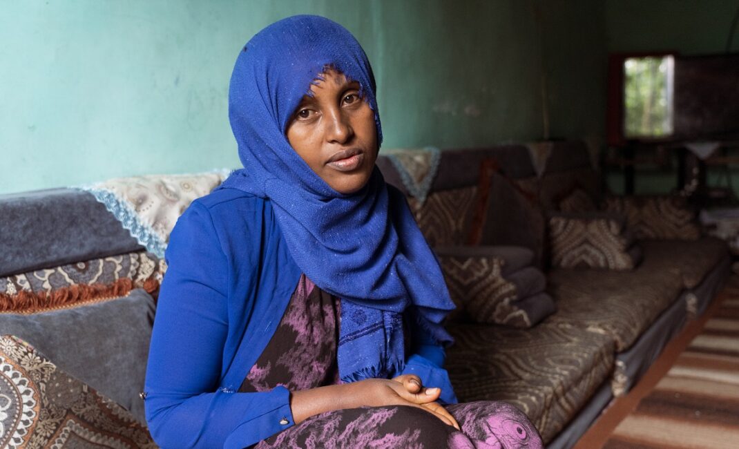 A woman wearing a bright blue hijab, blue blazer and burple dress sits on a couch in a cyan room. She is in her home in Addis Alem:  Photograph by Jaclynn Ashly for Lacuna story on religious tensions in Ethiopia.