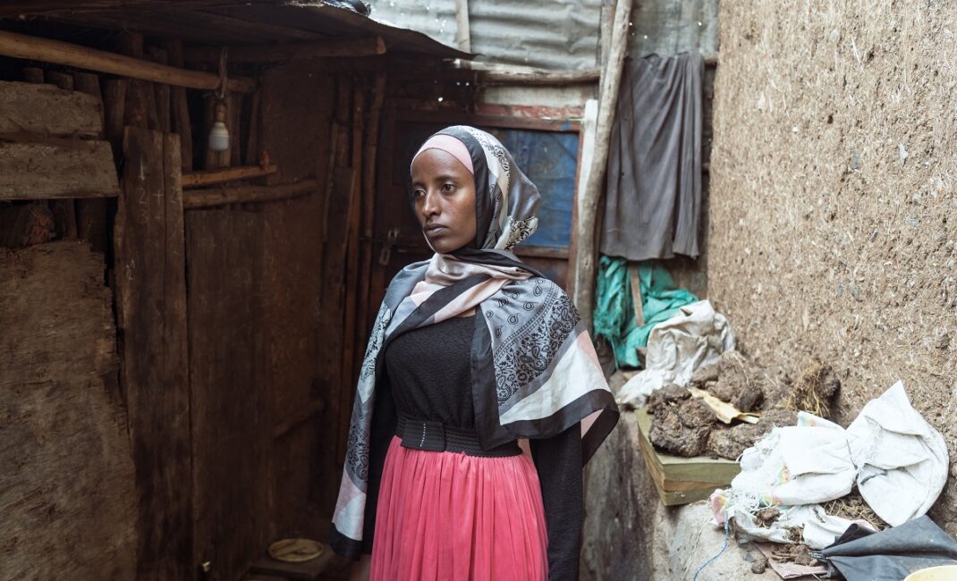 A woman in hijab and pink skirt stand in her village in Debark. Wooden and mud walls surround her with a singular light hanging:  Photograph by Jaclynn Ashly for Lacuna story on religious tensions in Ethiopia.